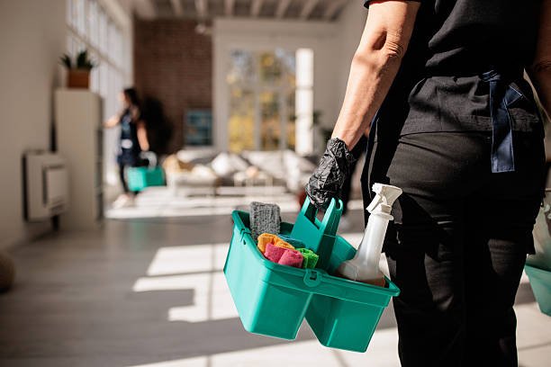 Dedicated cleaning professional carrying baskets of essential cleaning supplies for her colleague through a modern living space.