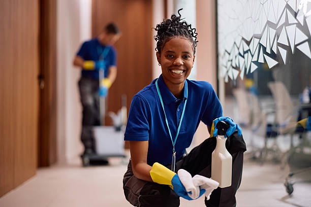 Happy African American female cleaner wiping glass walls in hallway of an office building and looking at camera.